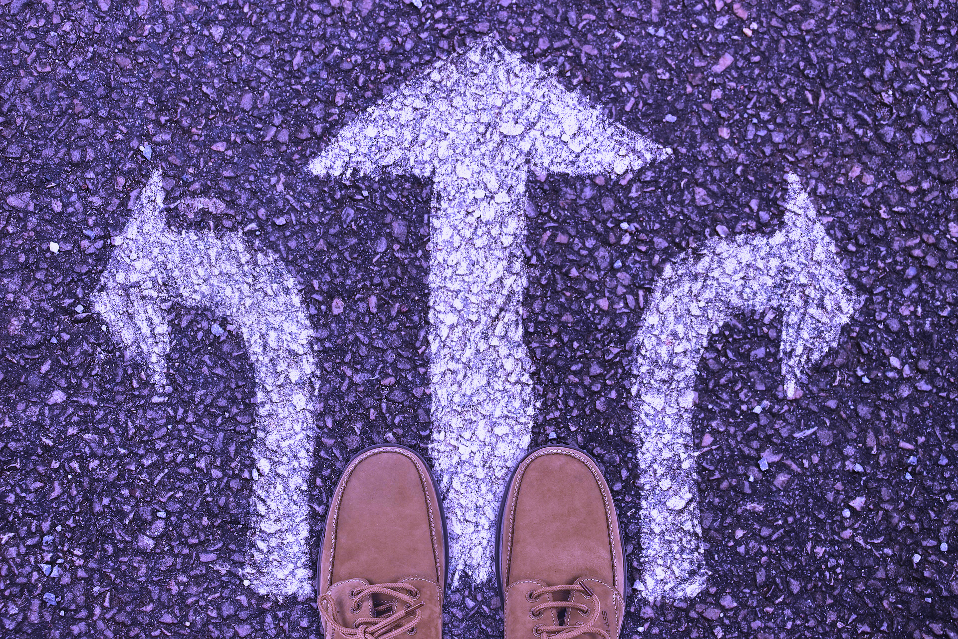 A pair of shoes stand in front of three diverging arrows painted on a road.